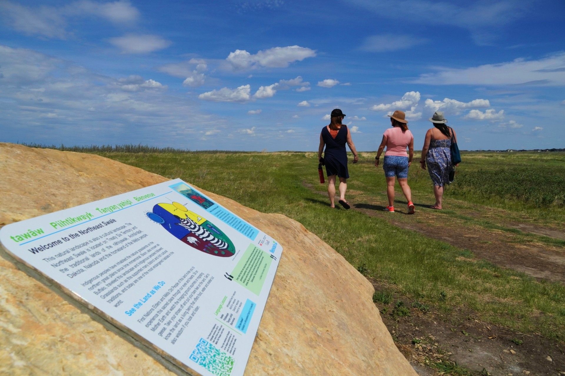 Three people walking ia a plain in the background. a park welcome sign at the forefront